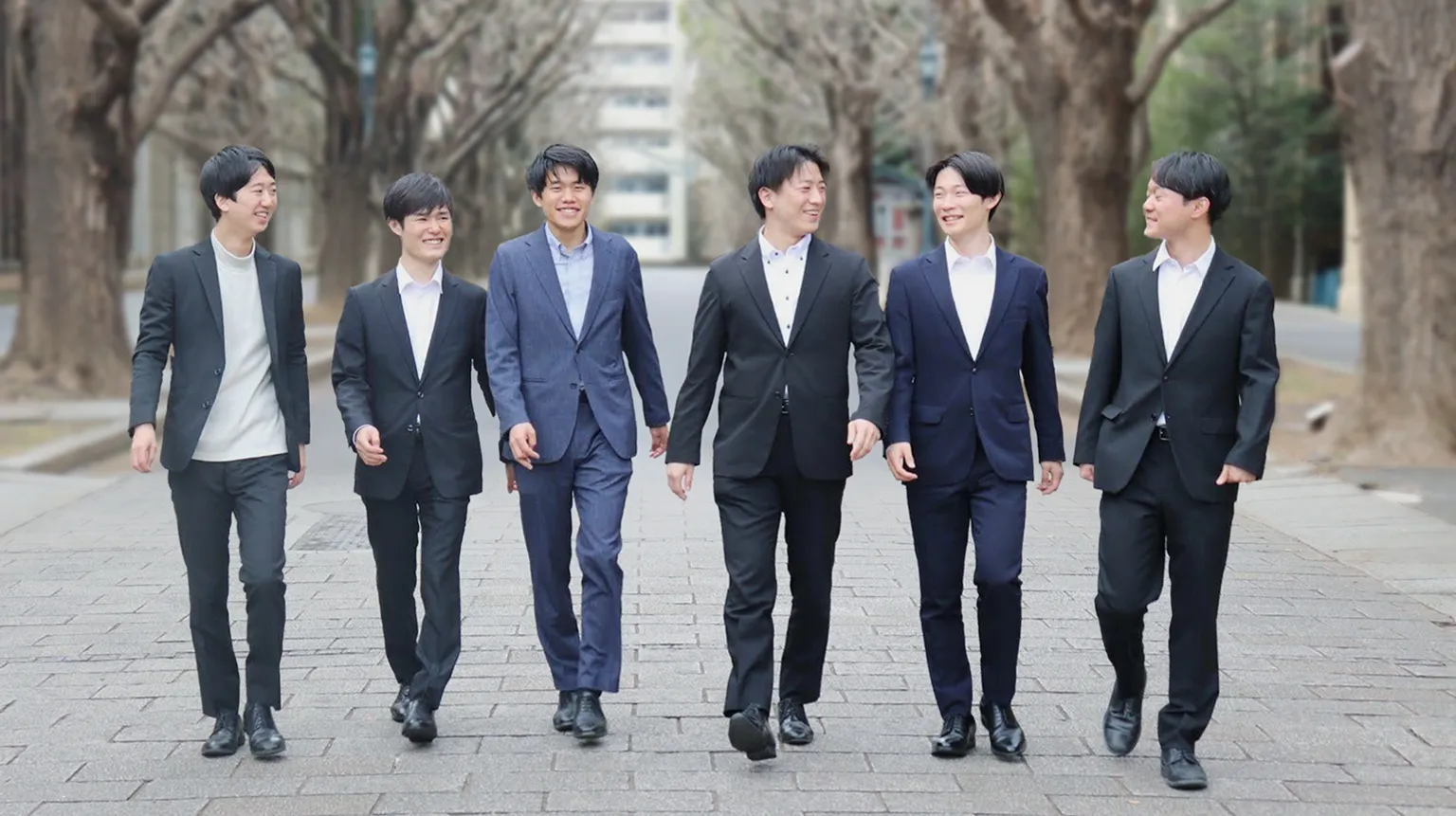並木道の中央を歩く、黒や紺のスーツを着用した6人の若い男性たちが、笑顔で談笑しながらこちらに向かって歩いてくる様子。