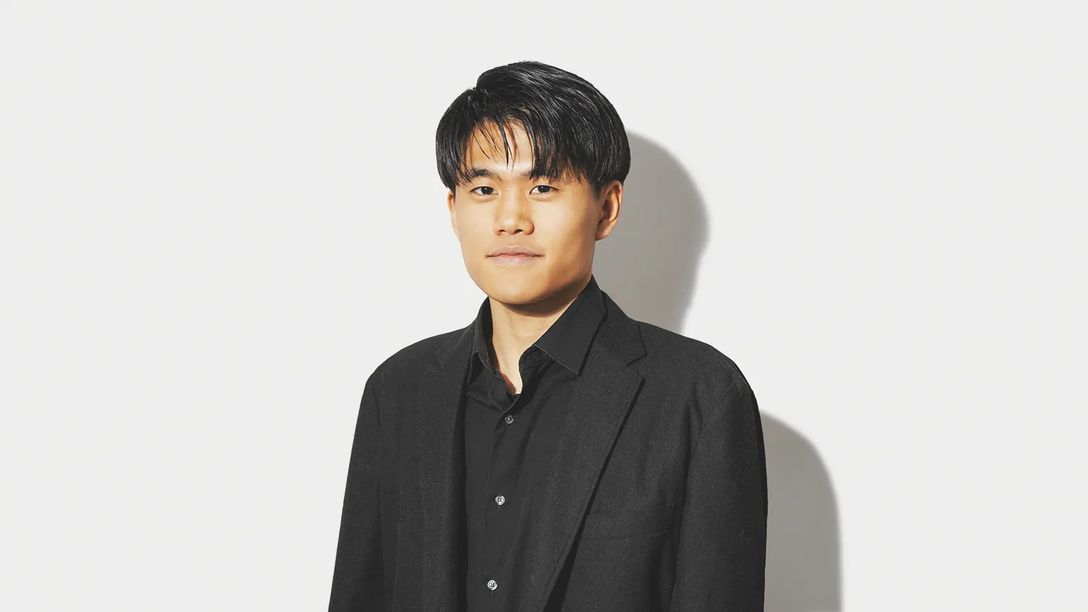 黒いシャツとジャケットを着用した若い男性のポートレートで、背景は白く、周囲に紫や黄色、グレーの幾何学的なグラデーションタイルやデジタルグリッドの要素が浮遊している。