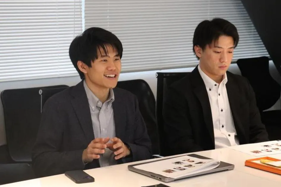 会議室で資料を前に座るスーツ姿の男性二人。左側の男性は笑顔で身振り手振りを交えて話し、右側の男性は視線を落として話を聞いている。