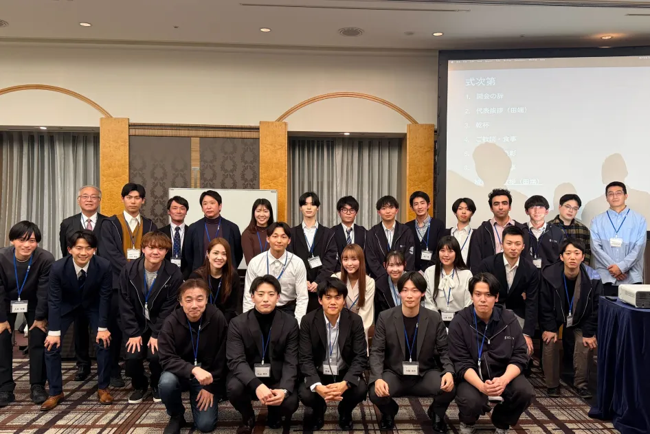 室内で「式次第」が投影されたスクリーンの前に集まる、スーツやカジュアルな服装の男女約25名の集合写真。